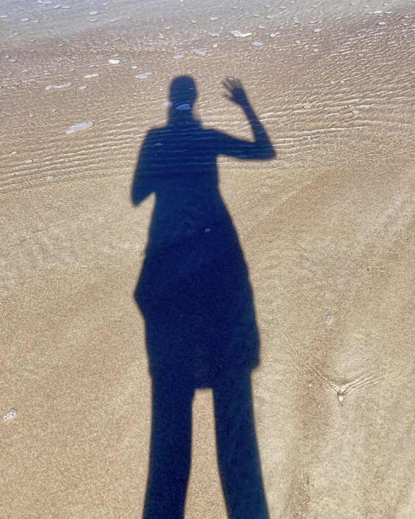 Photo of my shadow on sand and in shallow water. I am waving to the camera. 