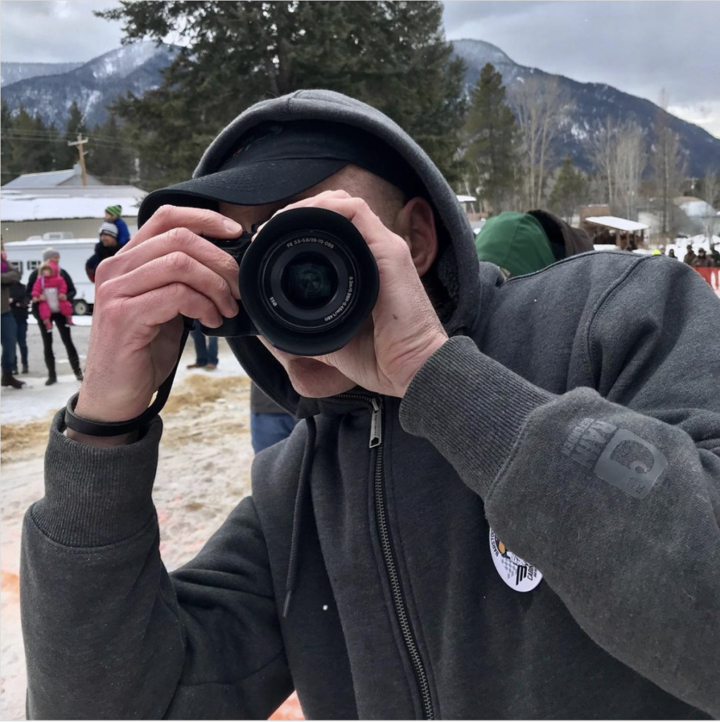 photo of a man with a black cap and grey sweatshirt looking at the photographer through the lens of a camera. 