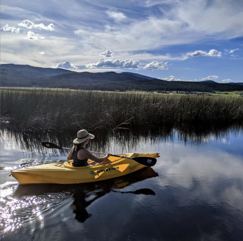 Christine in a yellow kayak on a river with tall grass and blue sky, with scattered clouds. 