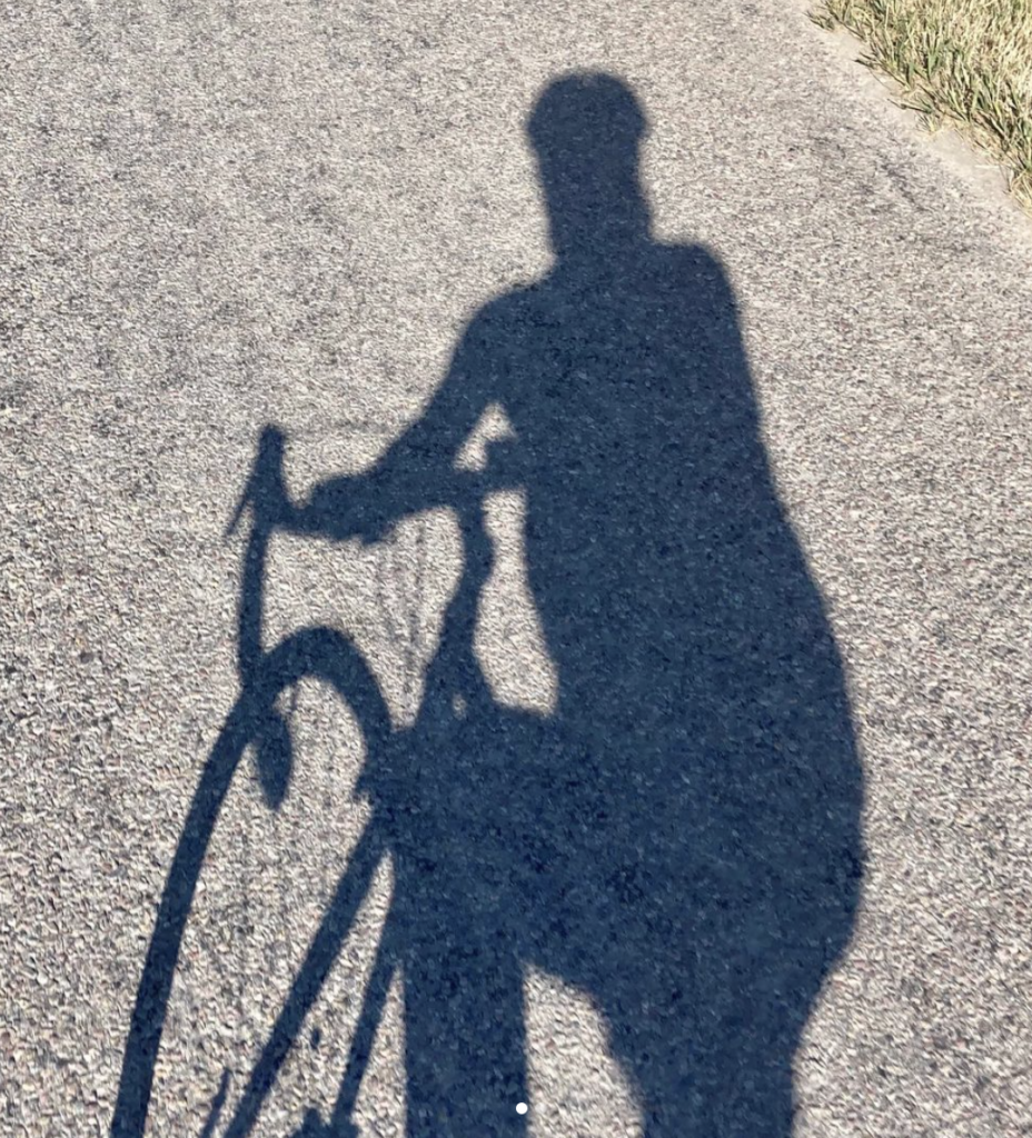 A photo of the ground with a shadow of Christine on a road bike, with a helmet. 