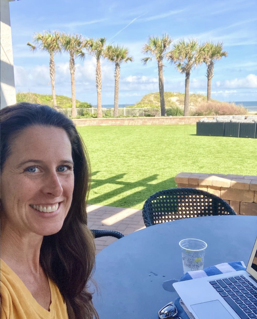 Photo of me smiling at the camera, with green grass, palm trees, dune, and the ocean in the near distance. In front of me is my laptop- not a bad place to work from, not bad at all. 