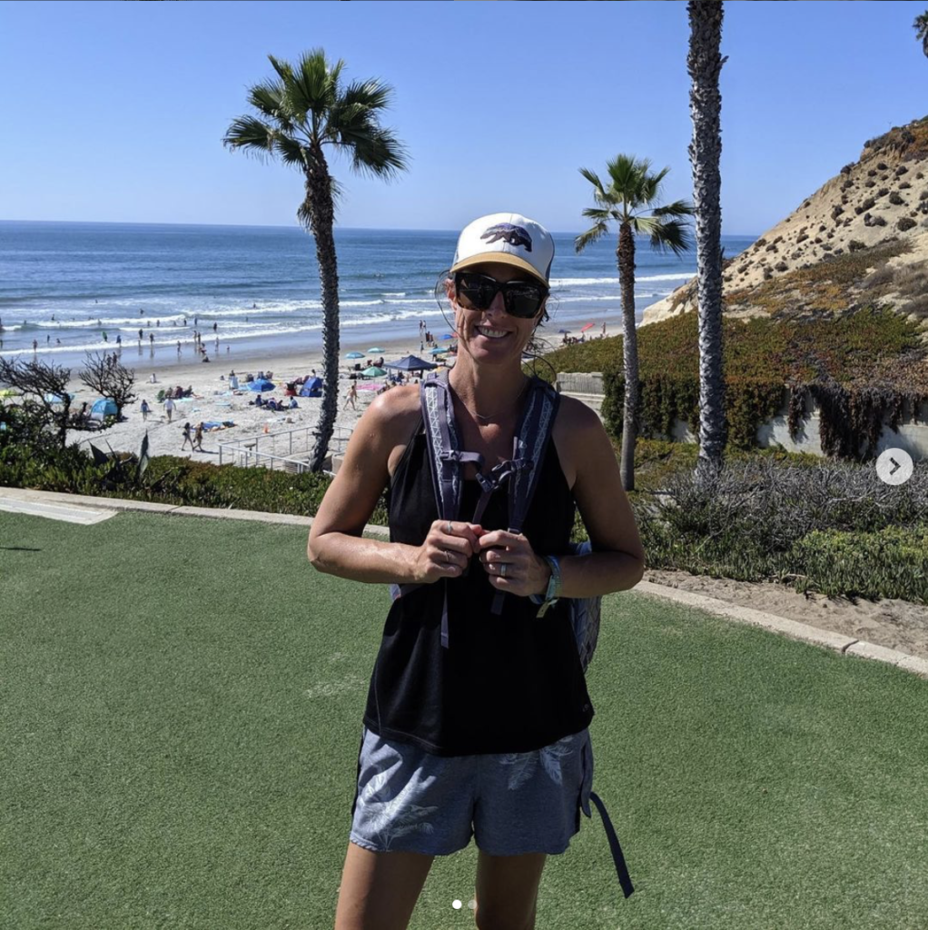 Christine in a baseball hat, tank top, shorts, with sunglasses, a backpack, and big smile at Solana Beach, CA, living her best life. 