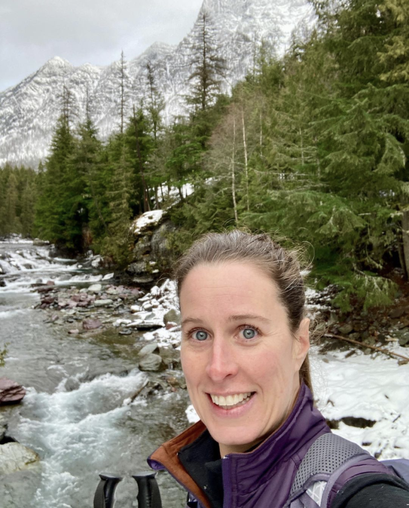 Photo of me, holding cross-country ski poles and standing on a bridge in Glacier National Park. In the background is a river, trees, and very large mountains. 