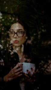 Woman holding a white coffee mug outdoors surrounded by dark green foliage, looking thoughtful.