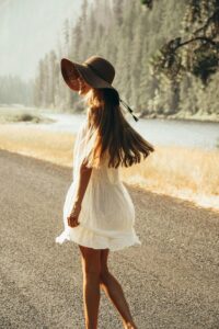 Woman in a white dress and sun hat walking down an open road with mountains and river in the background